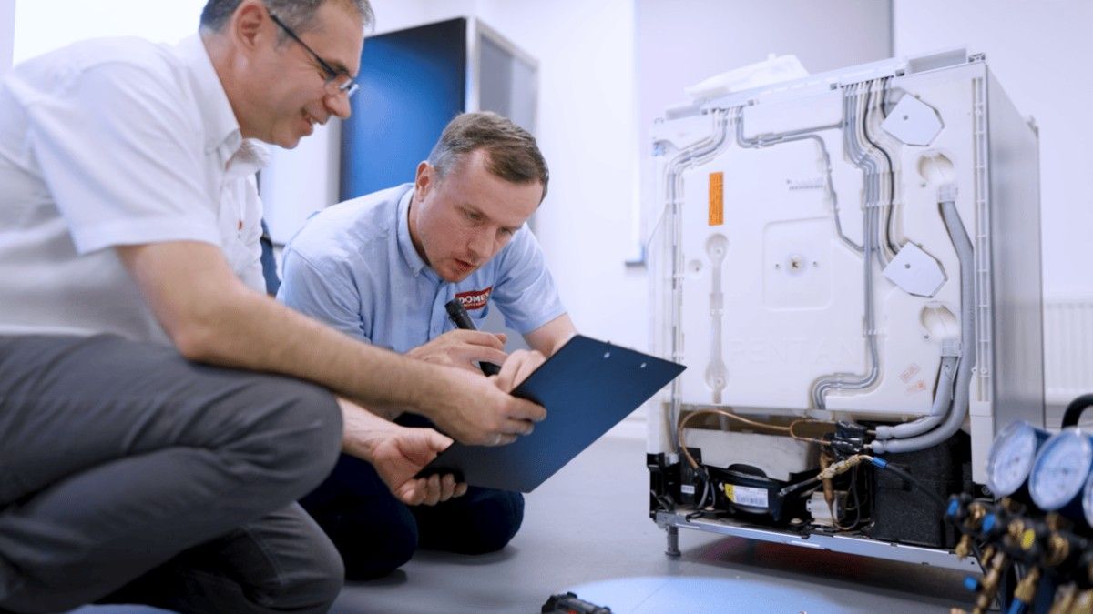 Two men kneel beside an open appliance, examining a clipboard and the exposed back panel, possibly performing a repair or inspection.