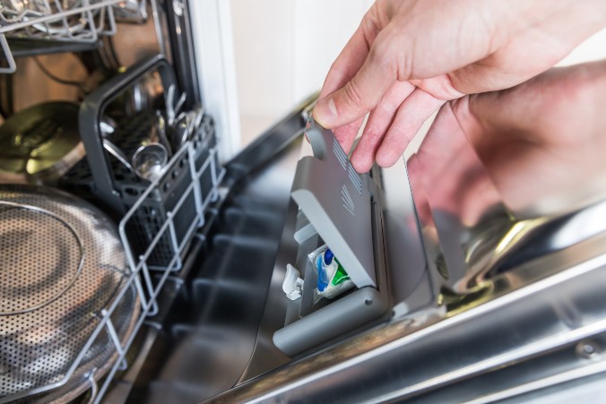 A person placing a detergent pod into the dispenser door of an open dishwasher.