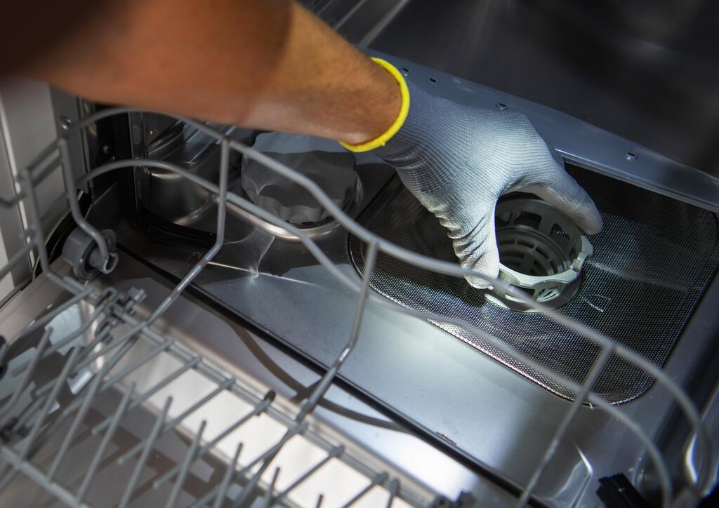 A gloved hand reaching into the bottom of an open dishwasher to remove the drain filter for cleaning. 