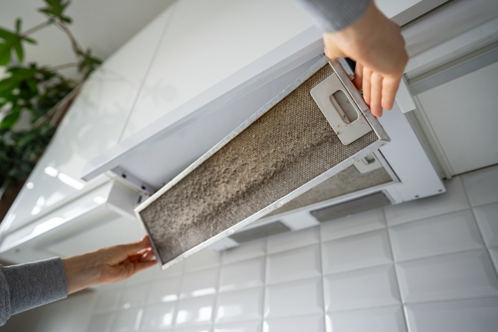 Close-up of hands pressing the clip mechanism to release a dirty metal filter from under a cooker hood. 
