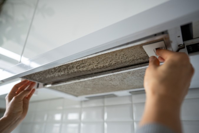 Person removing a grease-clogged metal mesh filter from a white kitchen extractor hood