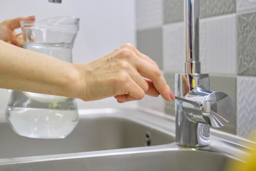 Hand turning on a kitchen tap to fill a glass pitcher for a water flow rate test.