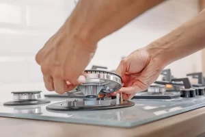 Hands lifting a burner off a gas hob, showing the gas jet, ignition electrode and flame safety probe during a safety check